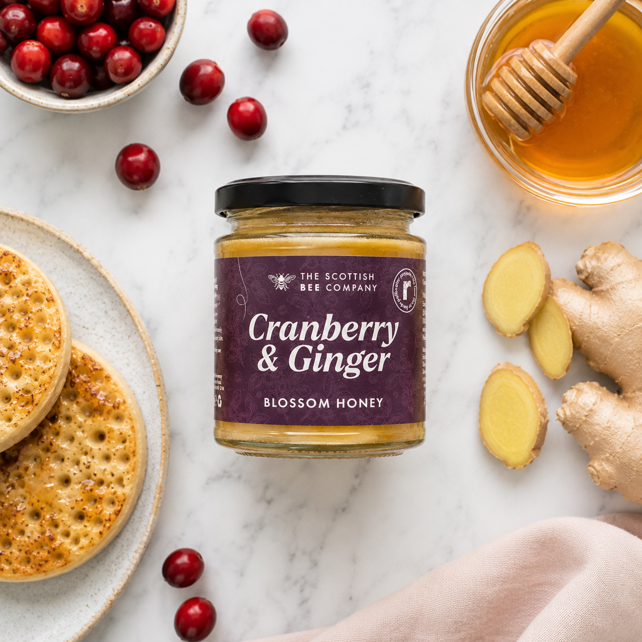 Jar of Cranberry & Ginger Blossom Honey from The Scottish Bee Company on a marble surface with crumpets, honey, and ginger.
